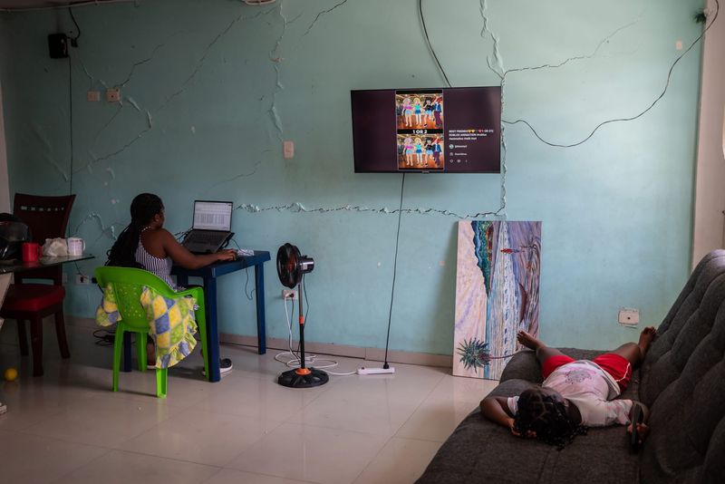 A woman works at a laptop beside a cracked wall in a Buenaventura home while a girl rests on the sofa, after the August 2026 earthquake
