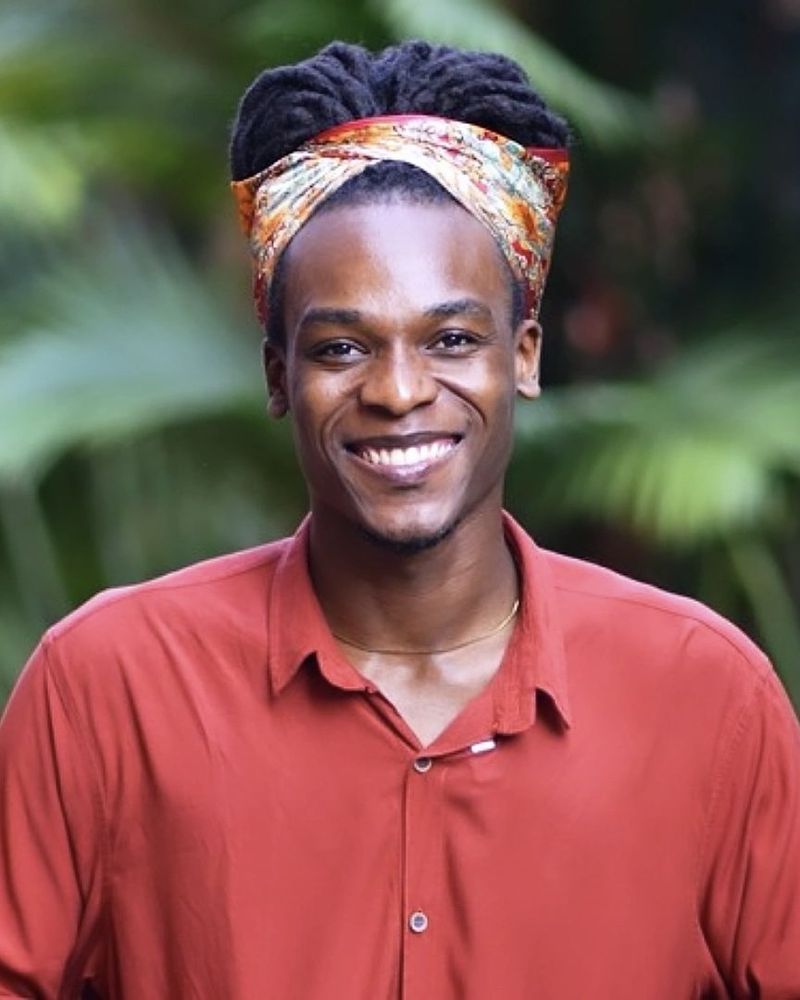 Deyner Caicedo Camacho, smiling, wearing a patterned headwrap and a rust-colored shirt, with tropical foliage behind him