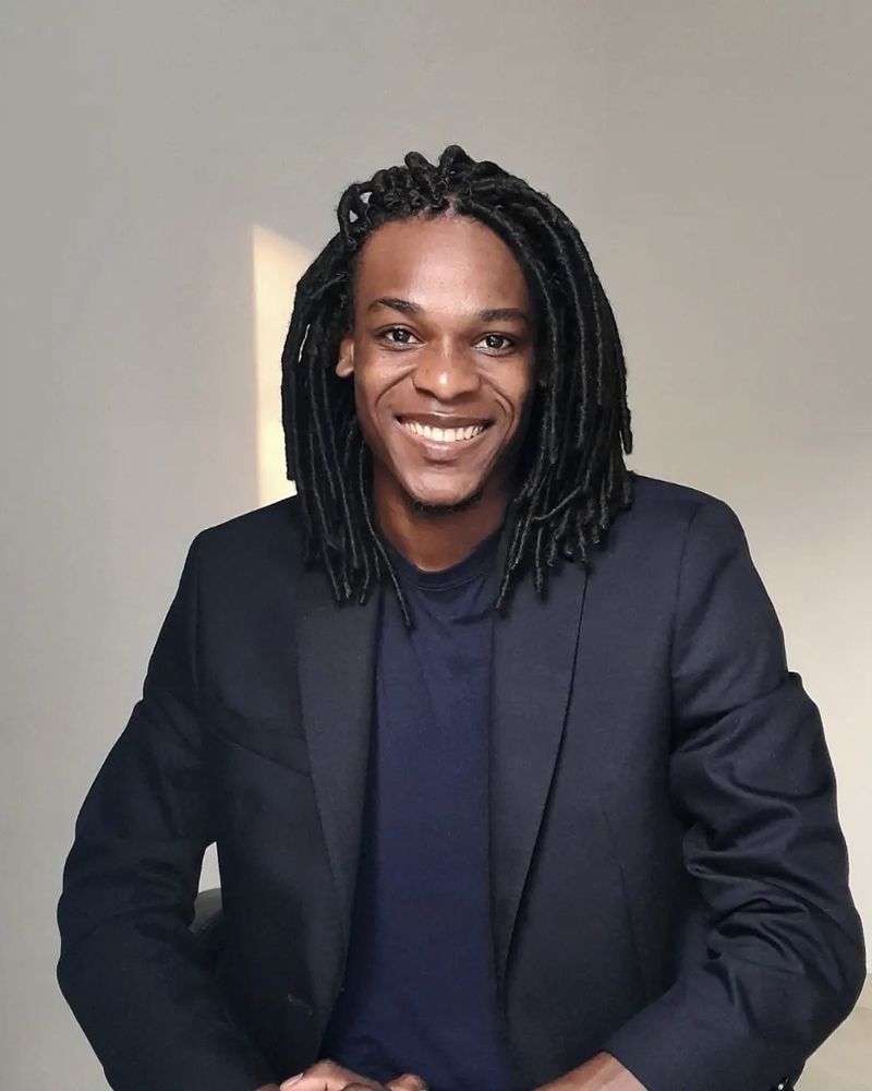 Deyner Caicedo Camacho, smiling, in a dark blazer against a pale grey wall